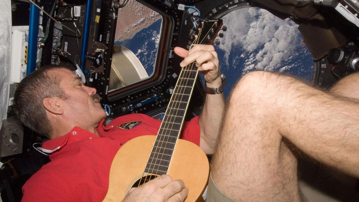  A man in a red shirt and shorts plays guitar while floating weightless in a cramped room full of windows. earth can be seen through the windows. 