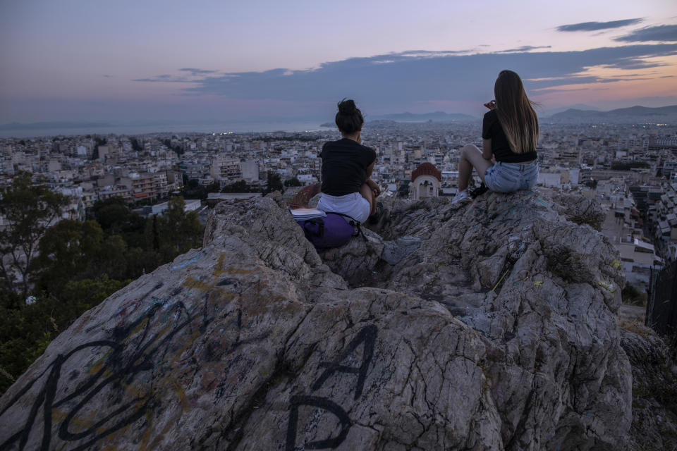 In this Thursday, July 4, 2019 photo, two young women sit on a rock covered with graffiti as they admire the view of Athens during a sunset. (AP Photo/Petros Giannakouris)
