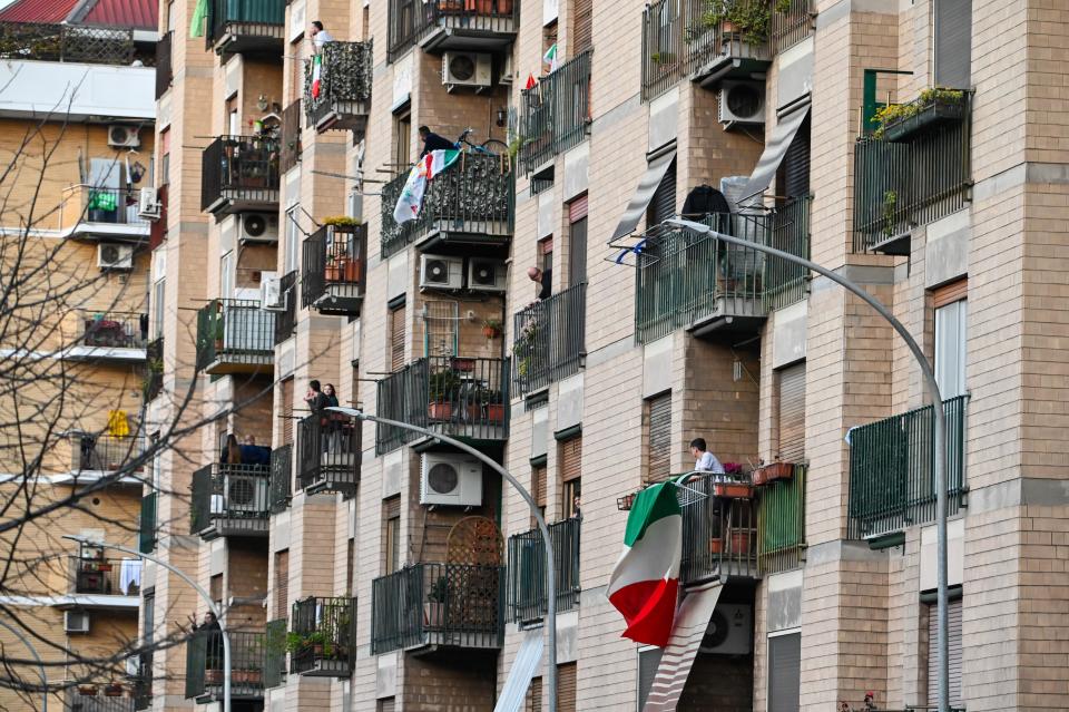 People wave and clap their hands next to a Italian flags, during a flash mob "Una canzone per l'Italia" (A song for Italy) at Magliana district in Rome on March 15, 2020. - Italy reacts with the solidarity of flash mobs circulating on social media to make people "gather" on balconies at certain hours, to play music or to get a round of applause. Italy on Sunday recorded 368 new deaths from the novel coronavirus, its highest one-day increase to date, taking the total to 1,809, the most outside China, official data showed. The number of infections has reached 24,747, a count released to the media by Italy's civil protection service said. The northern Lombardy region around Milan remained the European epicentre of the pandemic, officially reporting 1,218 deaths, or 67 percent of the Italian total. (Photo by Andreas SOLARO / AFP) (Photo by ANDREAS SOLARO/AFP via Getty Images)