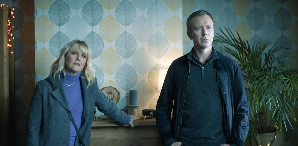 DI Ruth Calder (Ashley Jensen) and DC Sandy Wilson (Steven Robertson) question a suspect in their living room, as Calder leans on the mantlepiece