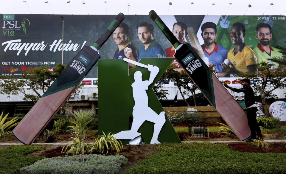 In this Saturday, Feb. 15, 2020 photo, a worker install a cut-out of huge bats next to a poster of cricketers displays along roadside in preparation of the upcoming Pakistan Super League, in Karachi, Pakistan. Security concerns stopped foreign cricketers from touring Pakistan four years ago when the country's premier domestic Twenty20 tournament was launched, forcing organizers to stage the event on neutral turf in the United Arab Emirates. When the 2020 edition of the PSL starts in Karachi on Thursday, Darren Sammy of the West Indies and Shane Watson of Australia will be among 36 foreign cricketers involved in the six franchises. (AP Photo/Fareed Khan)