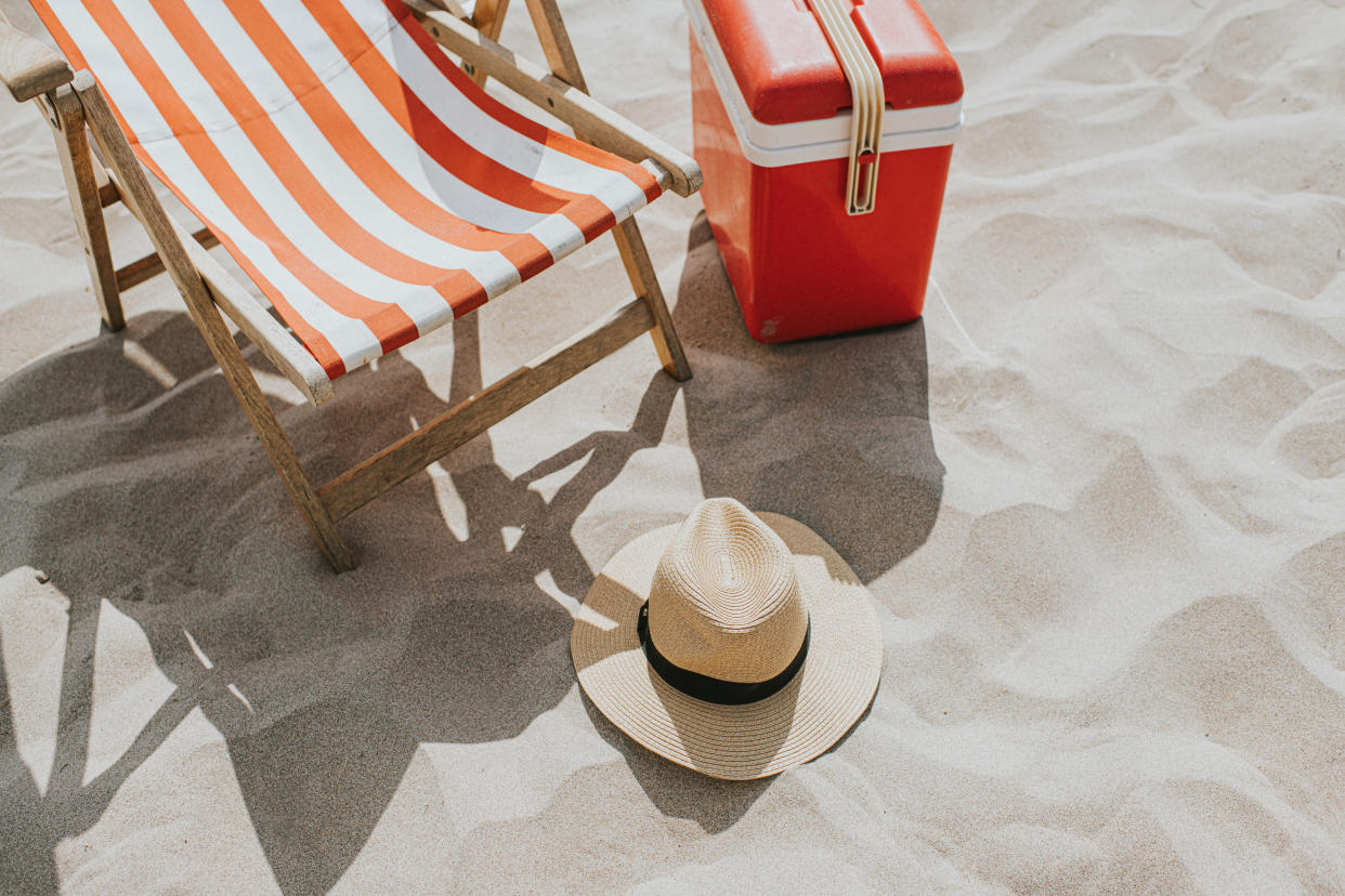 A relaxing coastal scene. A traditional style striped deckchair sits on a sandy beach. A sun hat rests on the ground and a cool box / cooler sits off to the side. Sand provides space for copy.