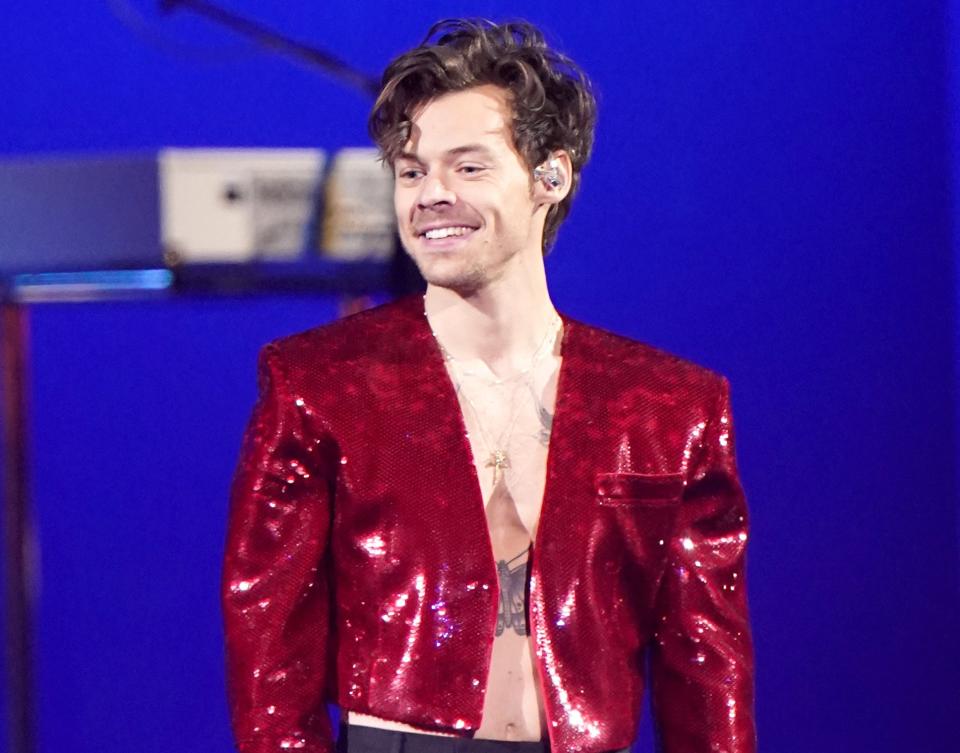 A close-up of Harry smiling in a shiny cropped jacket and bare-chested