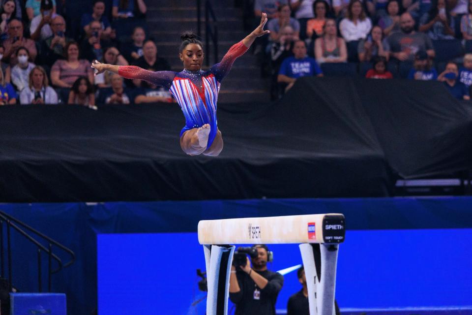 Simone Biles performs a split jump on the balance beam at a gymnastics competition, with a crowd watching in the background