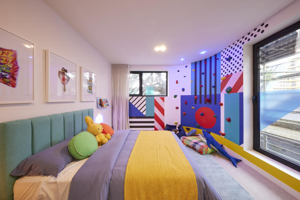 A wide shot of the room with a colourful double bed, a patterned curved wall with rock climbing on it and three pictures on the far left wall. 