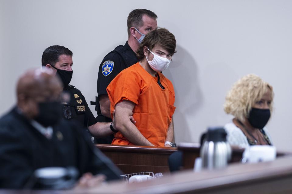 Officers flank a man in an orange jail outfit and mask