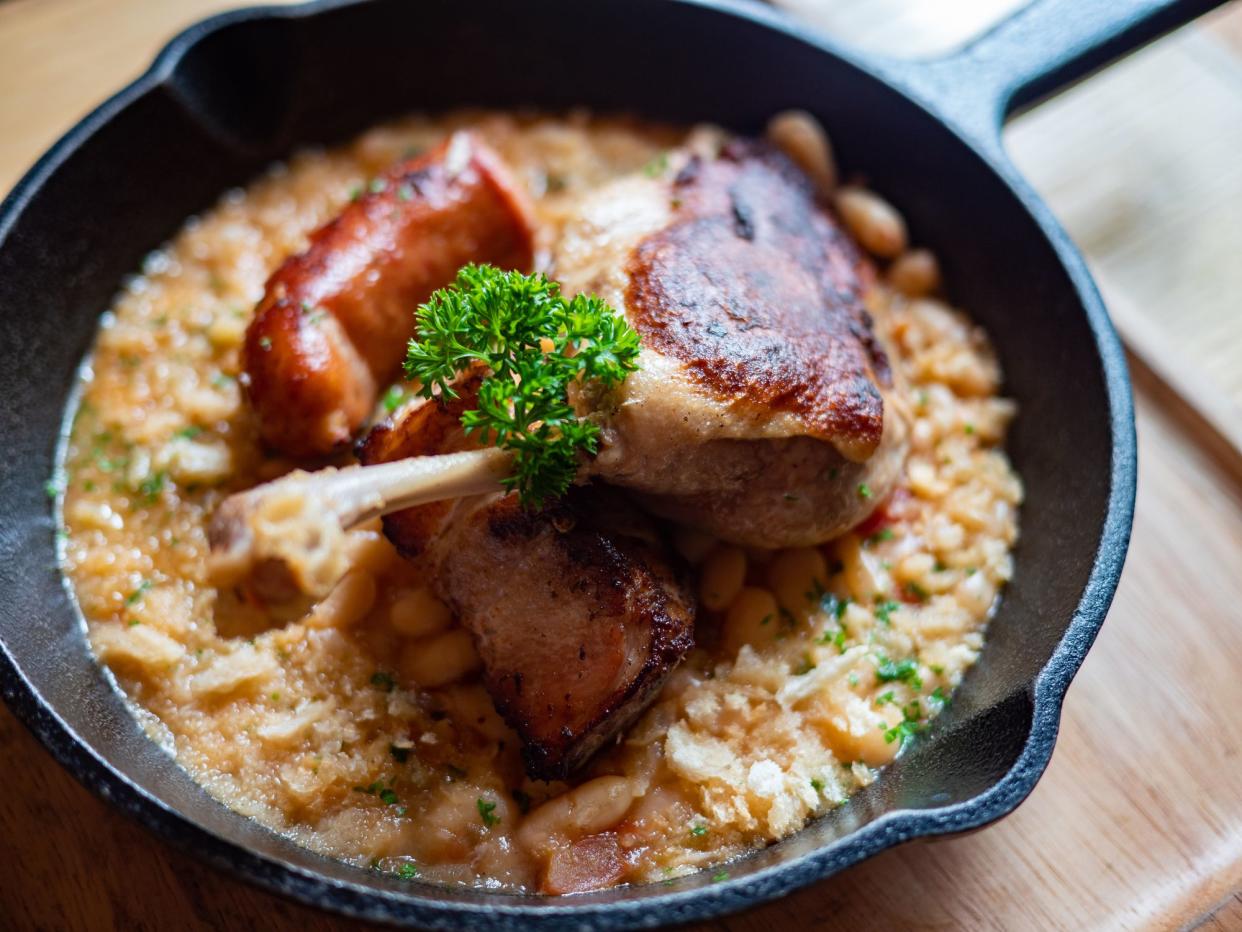 Sizzling hot, rich, slow-cooked Trio Cassoulet of duck confit, pork belly and smoked sausage on a bed of white beans in a black cast iron pan on a wooden table. Natural Light.