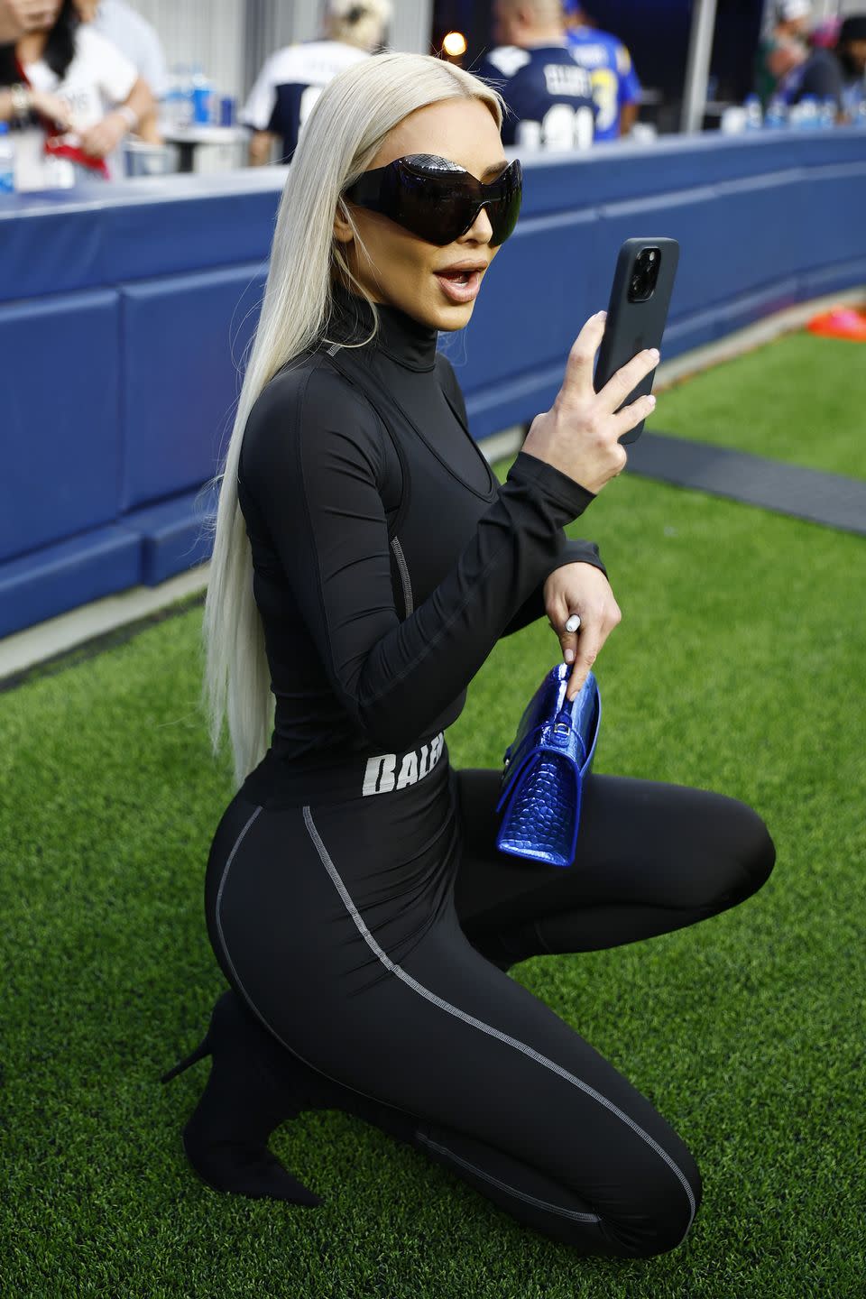 kim kardashian kneeling down and looking at her cell phone on a football field