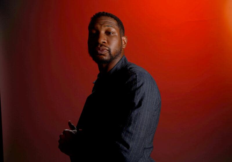 Jonathan Majors in a long-sleeve shirt posing at a side-profile against a dark red background with shadows in a corner