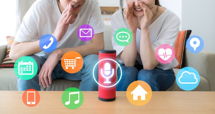 Icons for calls, music, groceries, and more float around a smart speaker to which two people are talking.