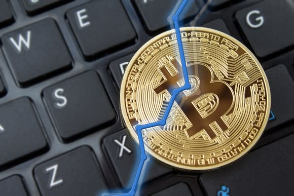 A rising stock chart overlaid on a physical gold bitcoin, with a keyboard in the background.