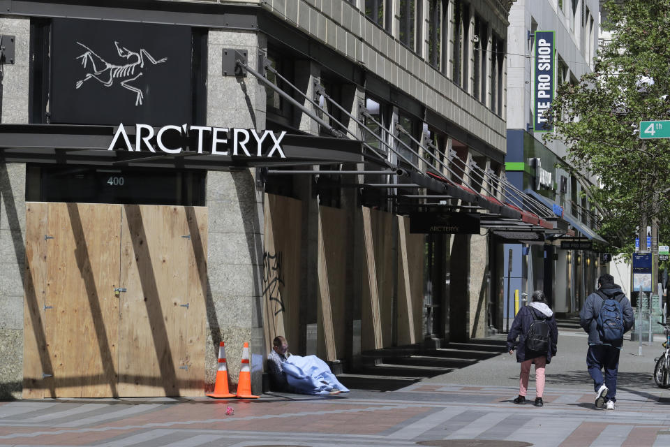In this May 6, 2020 photo, a person sits near a boarded up and closed Arc'teryx outdoor clothing store in downtown Seattle. Nearly all retail stores and restaurants in the area are currently closed or operating under reduced levels of service due to the outbreak of the coronavirus and state-wide stay-at-home orders, which has led to thousands of workers losing their jobs or being furloughed. (AP Photo/Ted S. Warren)
