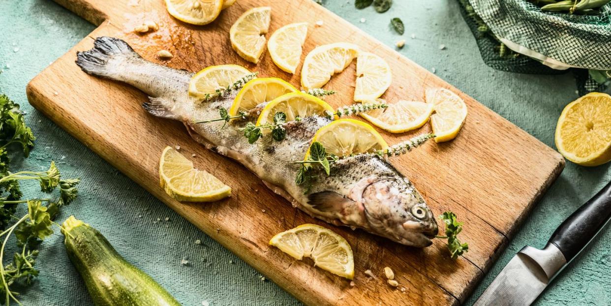 raw trout fish on cutting board stuffed with herbs and lemon slices on light blue kitchen table background with ingredients real cooking , step by step