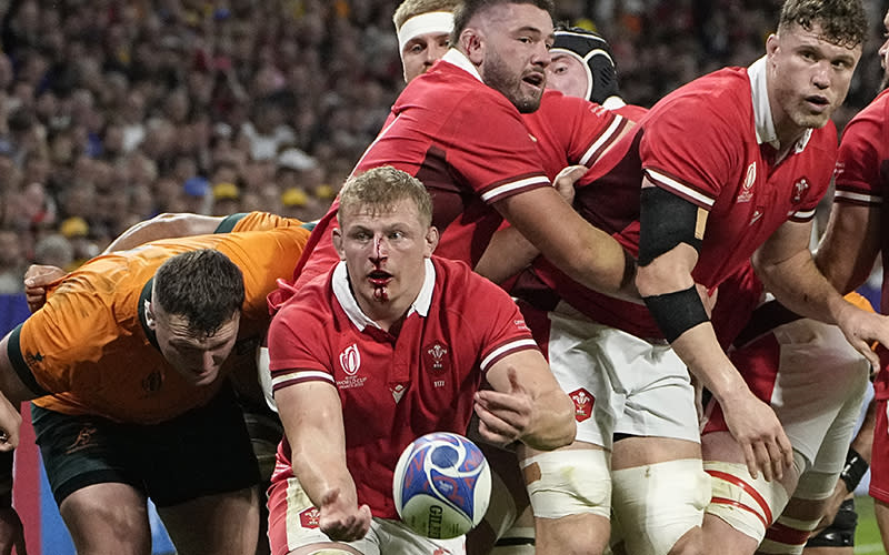 A group of rugby players are seen in the center. Some wear red jerseys and others wear orange once. crouched below the group is a player in a red shirt who has blood dripping down his nose and chin. He looks toward the viewer as he tosses the ball in that direction