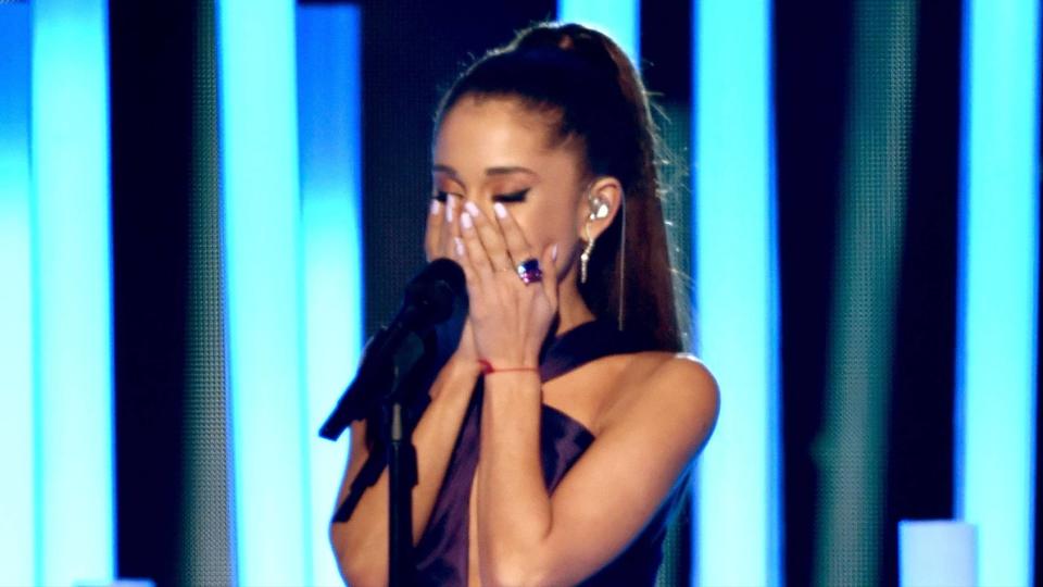 Ariana Grande on stage, with a microphone in front of her, covering her face with her eyes shut