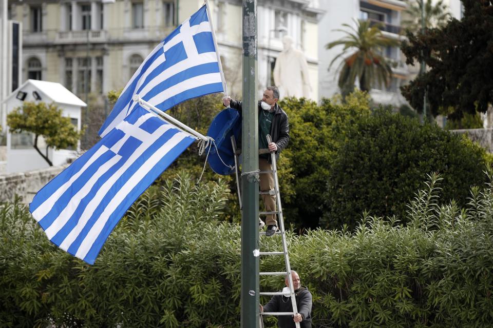 Municipal workers install Greek flags in front of the Greek parliament in Athens on Tuesday, March 24, 2020. An annual military parade has been canceled Wednesday amid a circulation ban imposed to slow the spread of the coronavirus but a flyover with jets and helicopters will take place. For some people the new COVID-19 coronavirus causes only mild or moderate symptoms, but for some it can cause severe illness including pneumonia. (AP Photo/Thanassis Stavrakis)
