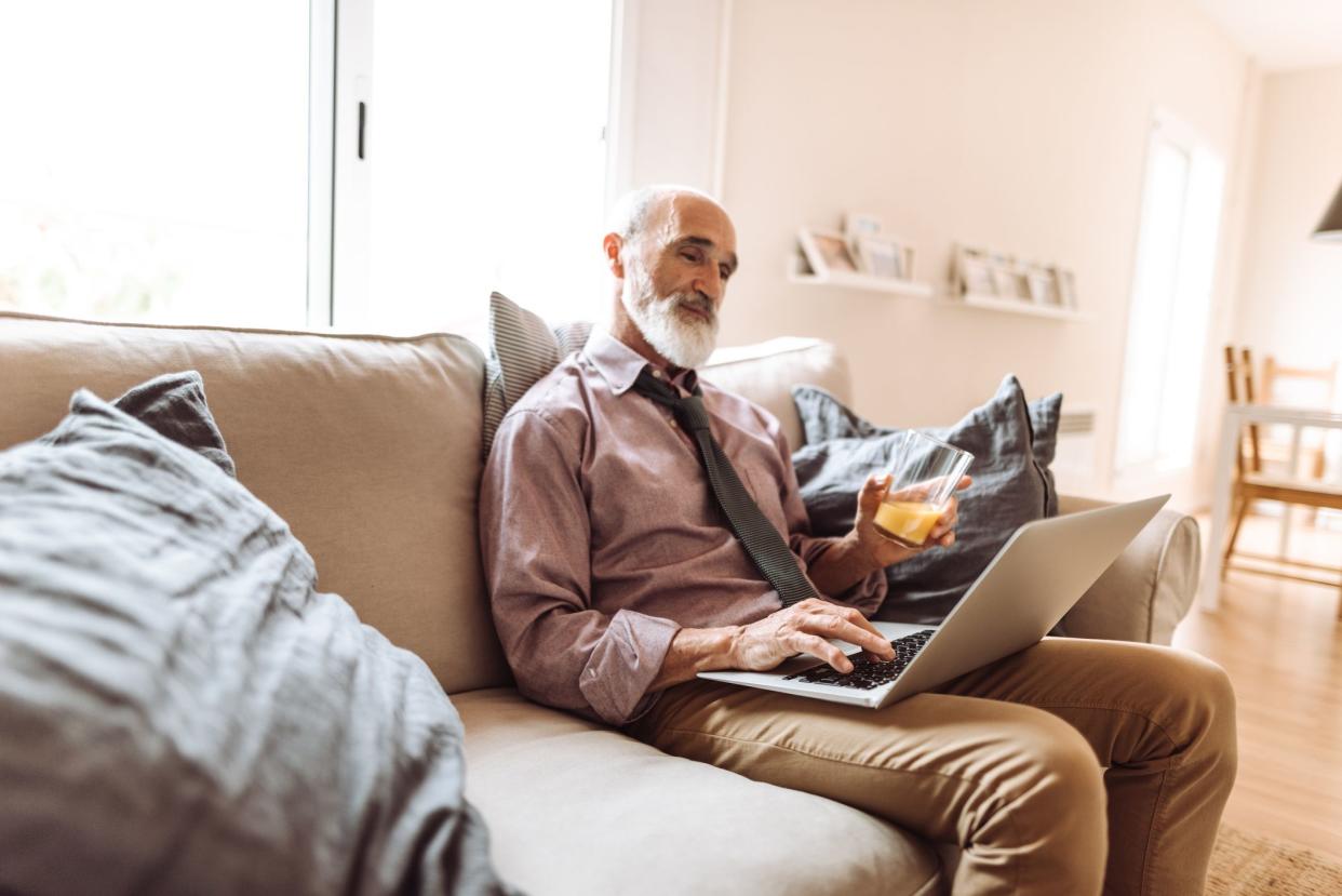 Relaxed senior man on a grey couch working with his laptop at home, holding a glass of orange juice, a blue throw pillow in the foreground, with another room in the background