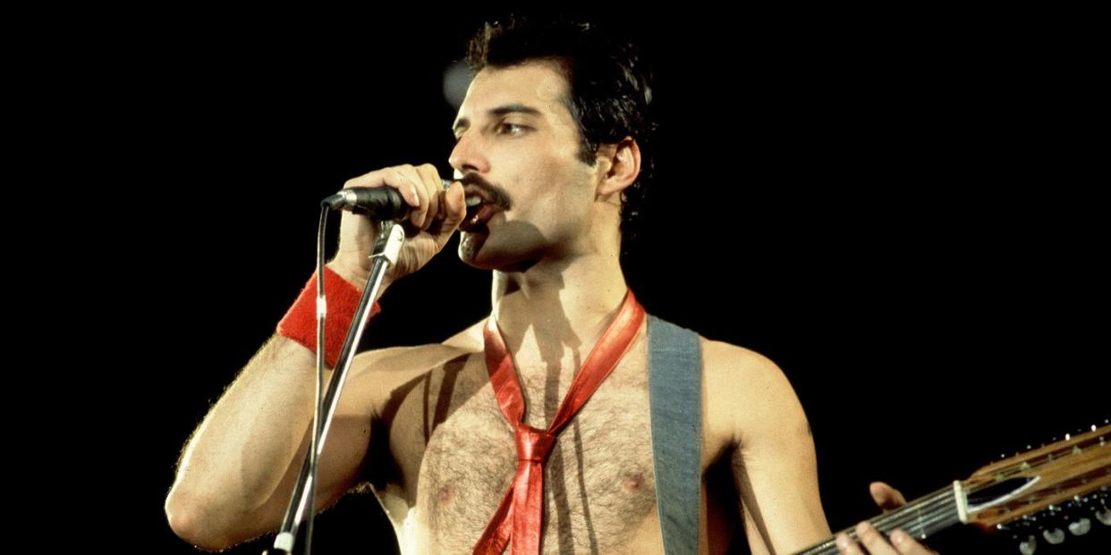 freddie mercury stands on a stage with a dark backdrop, speaking into a microphone on a stand, wearing no shirt, holding a guitar