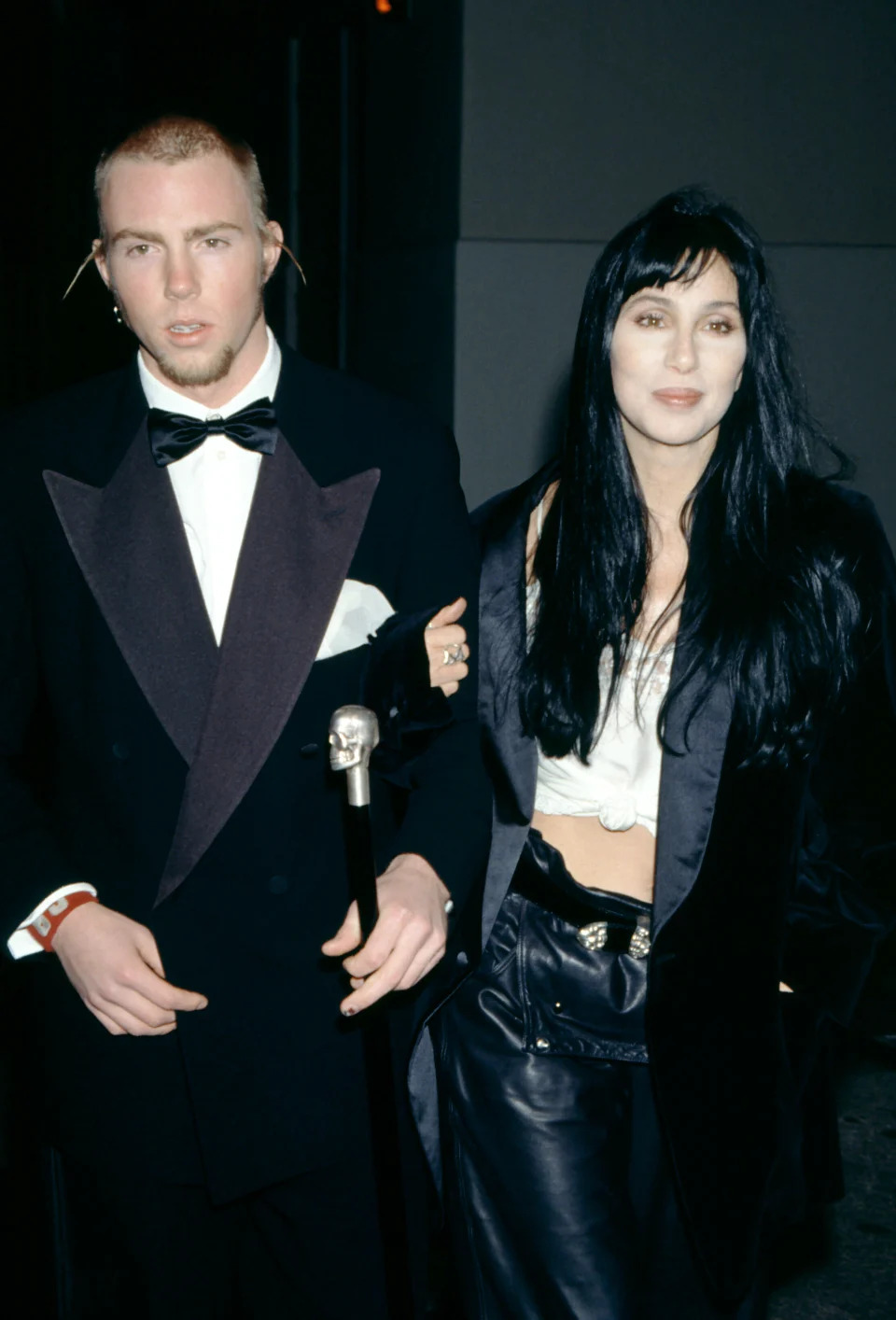 Cher, in a black leather outfit and black jacket, poses with her son, Elijah Blue Allman, in a black tuxedo holding a cane