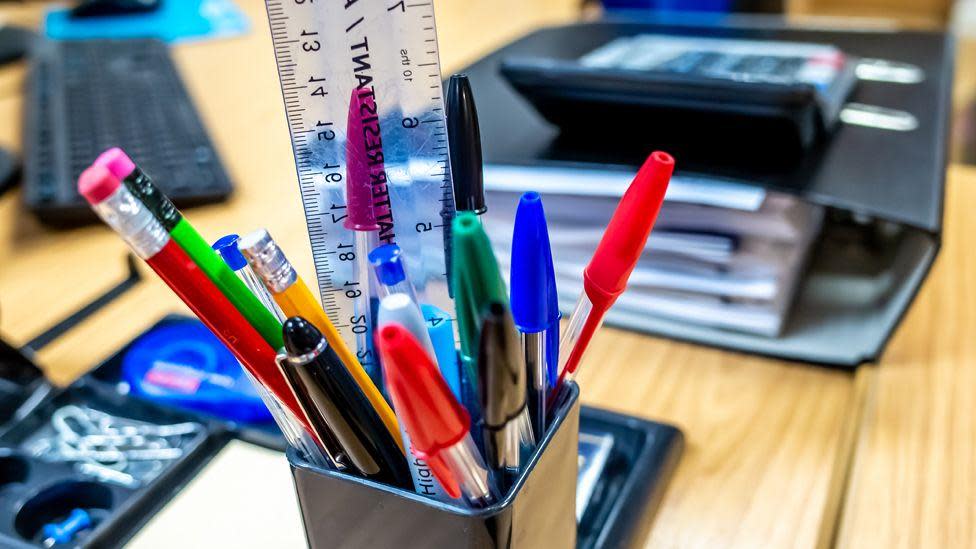 A desk containing a pot of pens, pencils and a ruler. In the background is a folder of paperwork and a computer keyboard.