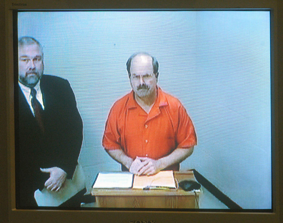 B2K stands before a camera at a podium with papers, with a man in a suit and tie standing next to him, in a setting appearing to be a courtroom