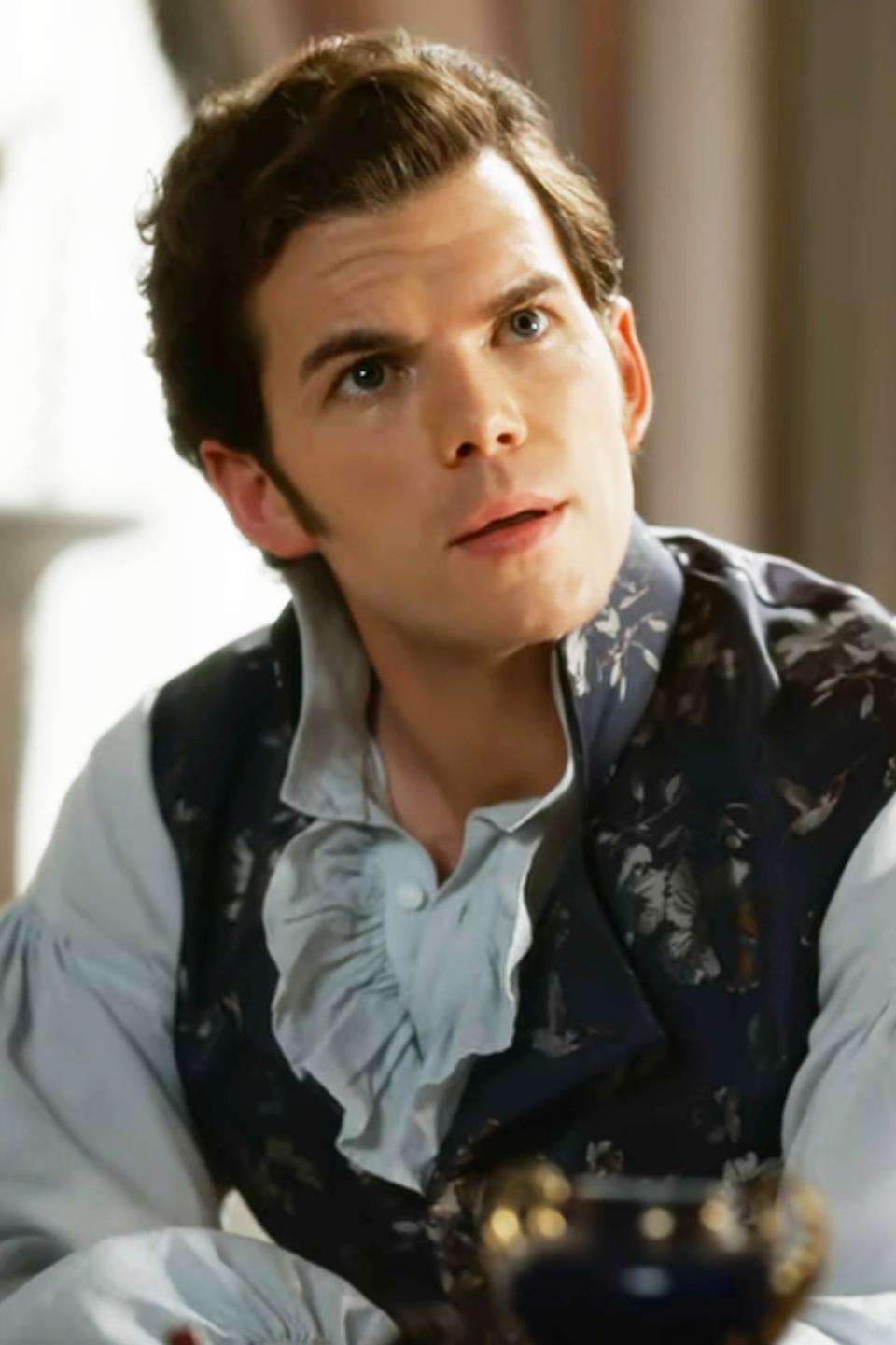 Luke Newton as Colin Bridgerton in a period costume with a ruffled shirt and vest, sitting at a table in a scene from 