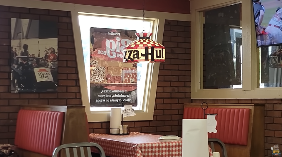 A Pizza Hut booth with red seating, a checkered table, and a stained glass style light fixture. Pizza Hut promotional posters are visible on the window