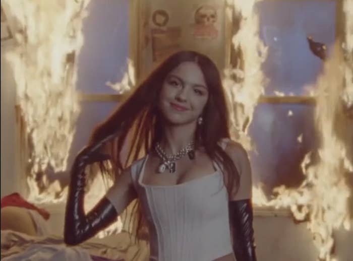 Olivia Rodrigo smiling in front of a room that's on fire