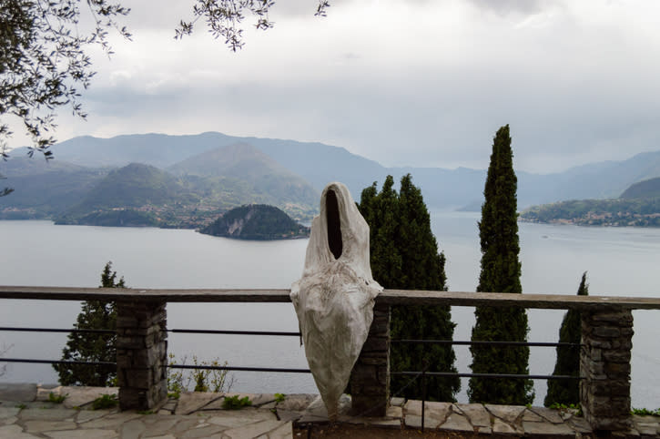 Sculpture resembling a cloaked figure without a face overlooks a tranquil lake and mountains