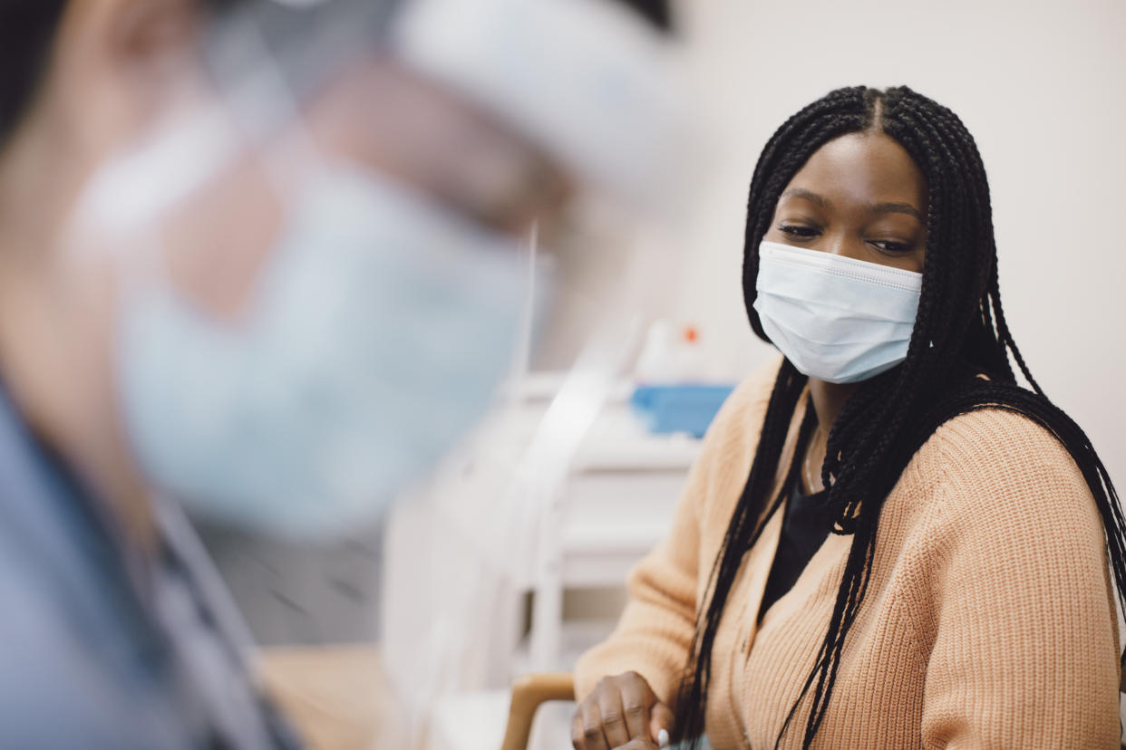 A Black woman wearing a mask is seated as a medical technician, in mask and visor, prepares a treatment.