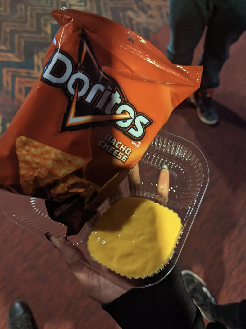 Person holding an open bag of Nacho Cheese Doritos with a container of cheese dip, standing indoors