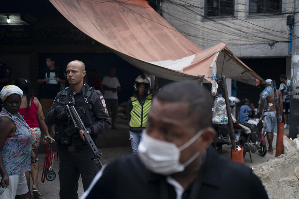 A police officer stands at the ready in a small market, as a local volunteer with a face mask walks away after distributing soap and detergent in an effort to avoid the spread the new coronavirus, in the Rocinha slum of Rio de Janeiro, Brazil, Tuesday, March 24, 2020. (AP Photo/Leo Correa)