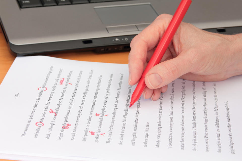 A hand holding a red pen is editing a printed document with corrections marked in red ink, placed on a desk next to a laptop