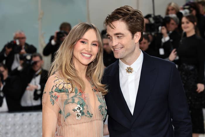 Suki Waterhouse and Robert Pattinson pose at a formal event. Suki wears a sheer dress with floral embroidery, and Robert wears a dark suit with a brooch