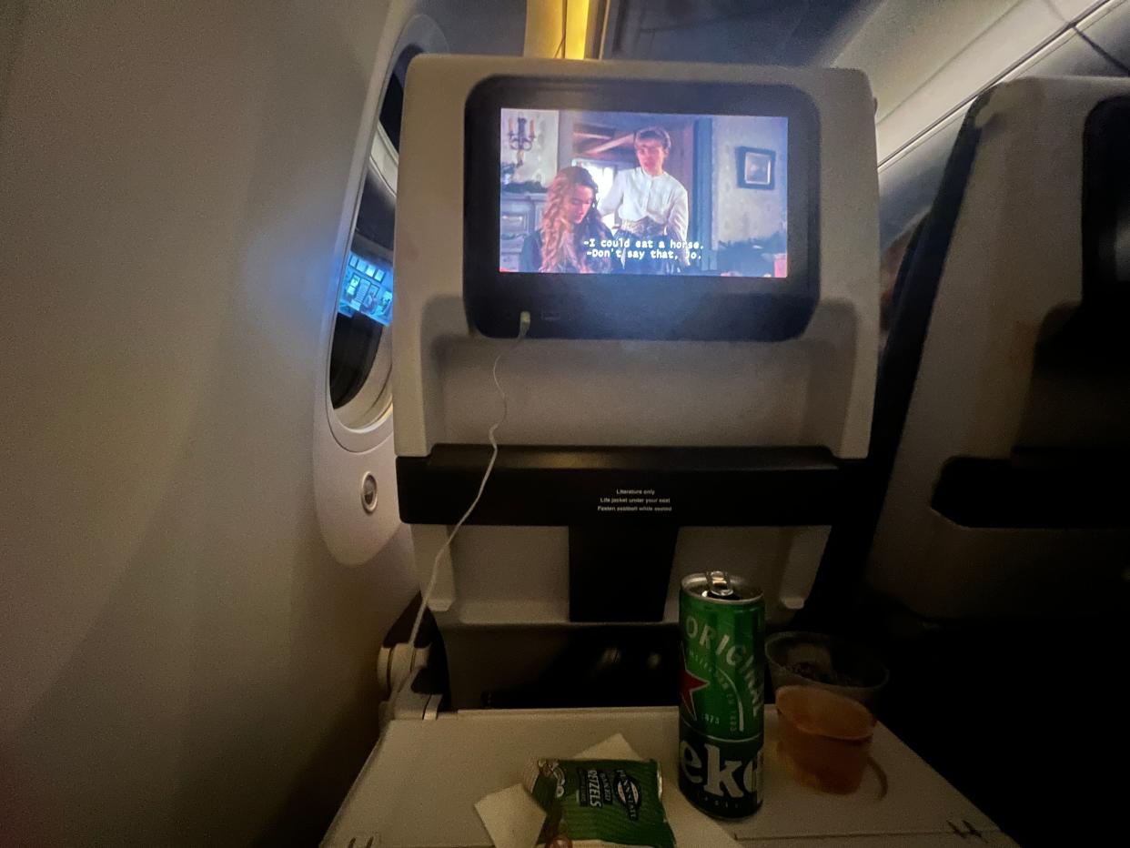 A first-person view of an economy class seat on a British Airways Boeing 787, with a movie on the screen, and Heineken a beer and pretzels on the tray table