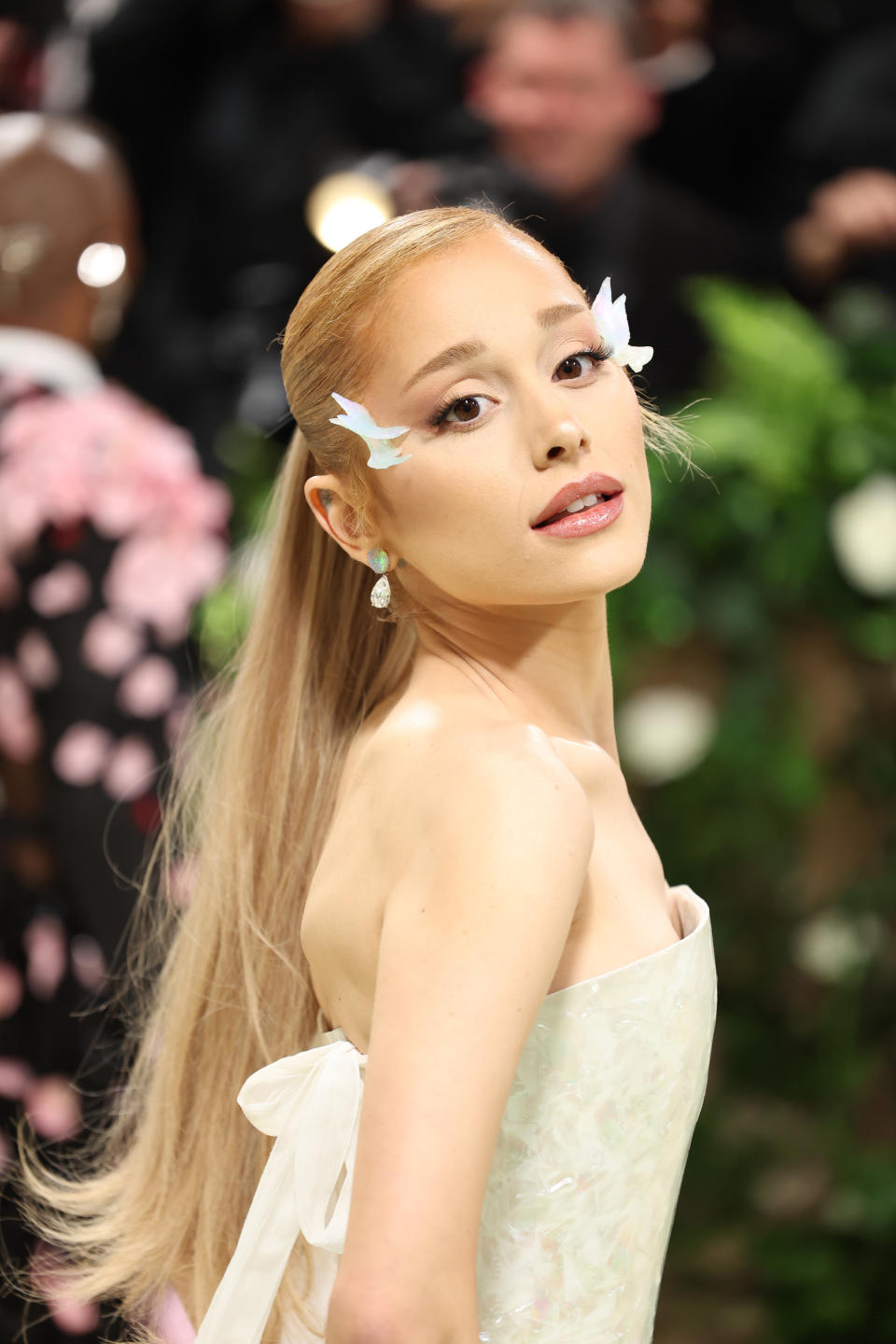 Ariana Grande on the red carpet, wearing a strapless gown with intricate detailing, featuring white butterfly accessories in her hair