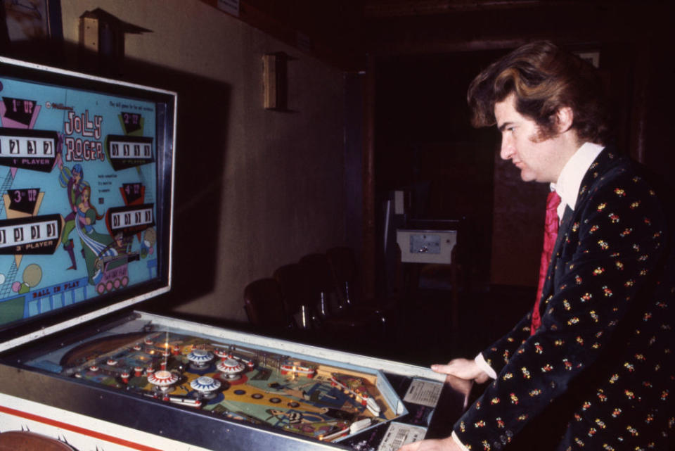 A man in a stylish suit with a patterned jacket and pink tie playing a vintage pinball machine named "Jolly Poker" in a dimly lit arcade