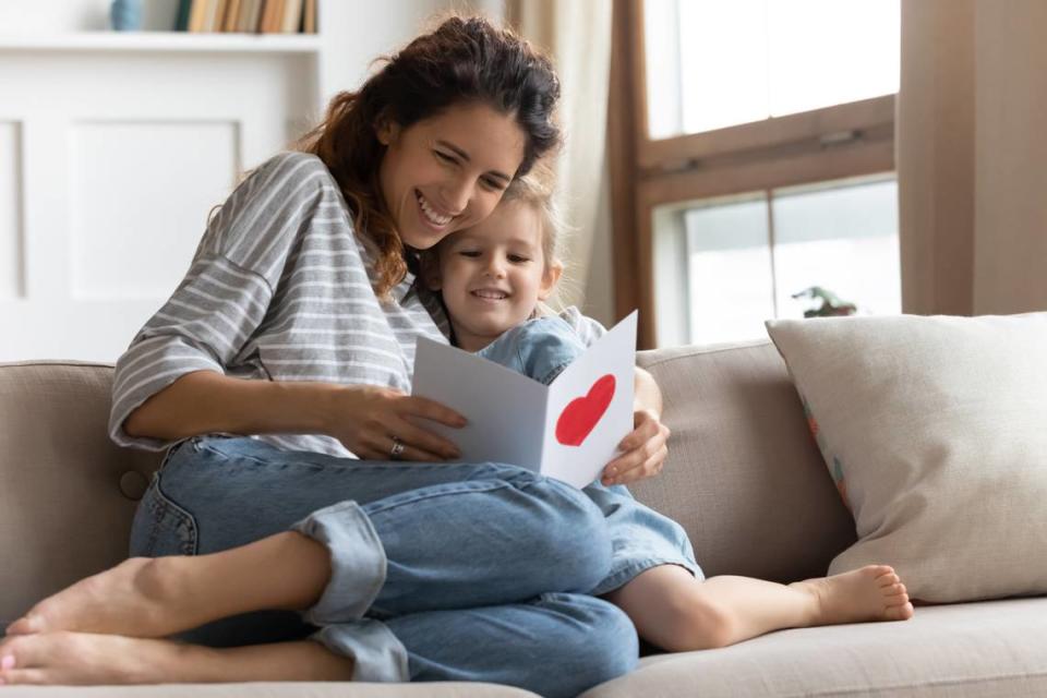 Happy young curly mother relaxing on sofa, embracing small preschool daughter, reading congratulations wishes in birthday card, feeling proud. Excited mommy enjoying greetings in postcard with child.