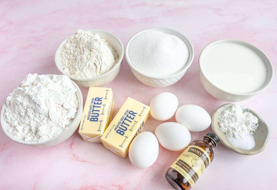 all of the ingredients for the recipe in bowls, including flour and sugar with eggs and sticks of butter on a pink background