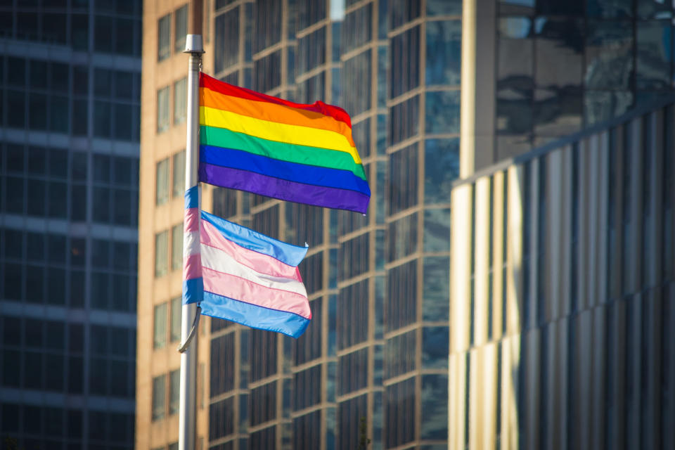 Pride and Trans Flag waving in the wind in a downtown urban setting.  Office buildings can be seen behind.