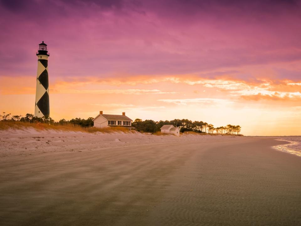 A view of the Cape Lookout lighthouse just before sunset. The clouds on the top of the frame are a soft fuchsia, with the sky underneath them a stark creamsicle orange, the lighthouse off to the left of the screen is checkered white and black.