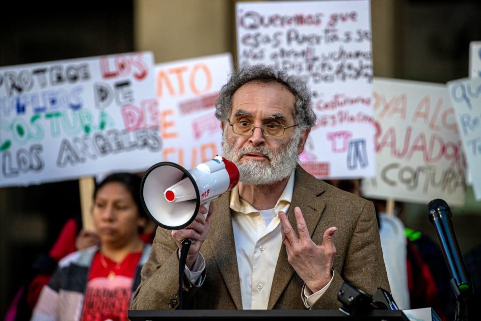 A man speaks into a megaphone at a rally as people behind him hold signs.