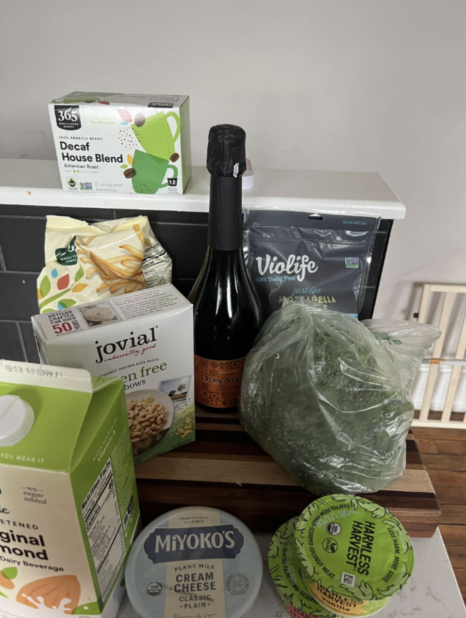 Various grocery items including decaf coffee, pasta, champagne, broccoli, garlic spread, almond milk, cream cheese, and Violife cheese are placed on a kitchen counter