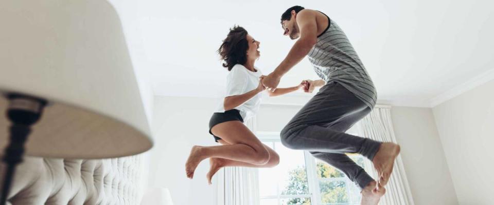 Man and woman holding hands and jumping together on bed. Couple having fun in bedroom.