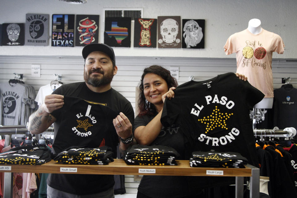 In this Thursday, May 21, 2020 photo, artists Charlie Monarrez, 46, and Monica Monarrez, 36, hold up "El Paso Strong" clothing at the print shop where they work in El Paso, Texas. Charlie designed the print after the Aug. 3 mass shooting using a slogan that united the city. Monica said she worked so hard printing the shirts and selling them to raise money for the shooting's dozens of victims that she was hospitalized with exhaustion after two weeks. (AP Photo/Cedar Attanasio)