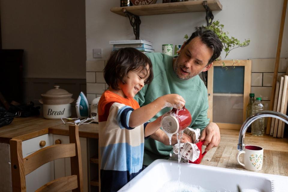 A waist-up shot of a male child and his dad washing and drying dishes together in their home. They are both smiling and wearing casual clothing.