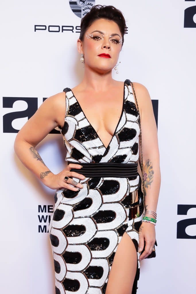 Em posing on the red carpet in a patterned dress with a deep V-neck and thigh-high slit, with intricate eye makeup and tattoos on one arm