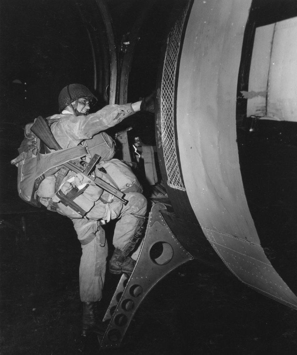 A heavily burdened paratrooper, armed with a Thompson M1 submachine gun, climbs into a transport plane bound for France