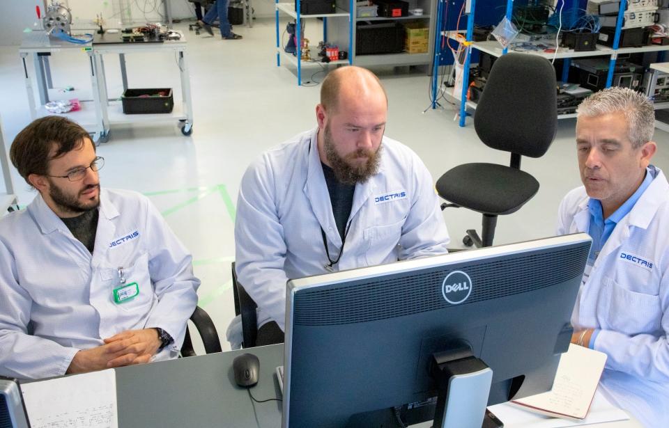 Three men in white lab coats sit at a desk looking at a computer