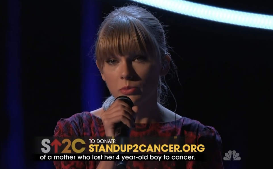 Woman on stage holding microphone with "StandUp2Cancer.org" text and donation information on screen